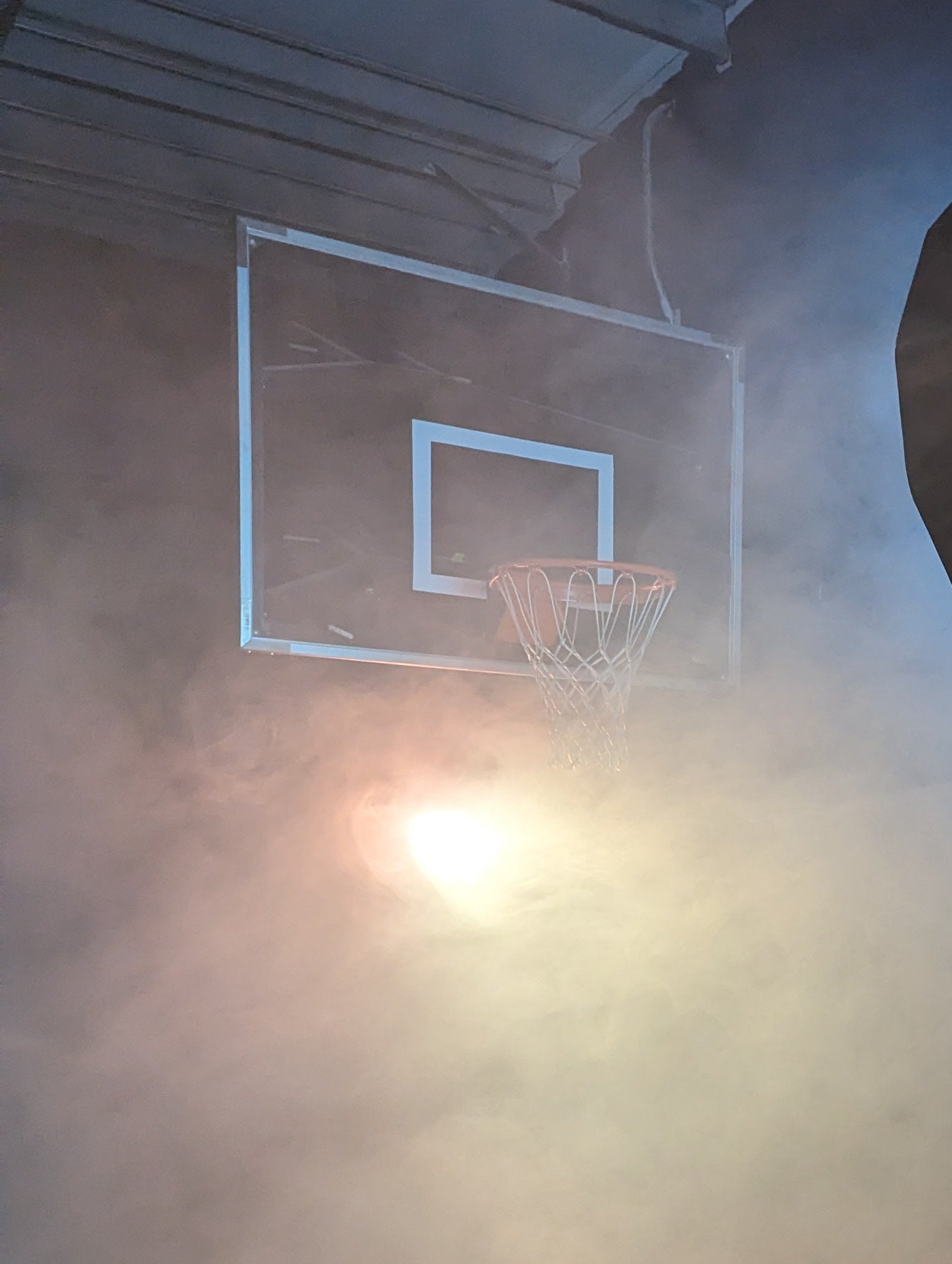 Basketball hoop with a large reflector and camera on a stand in a dimly lit indoor setting.